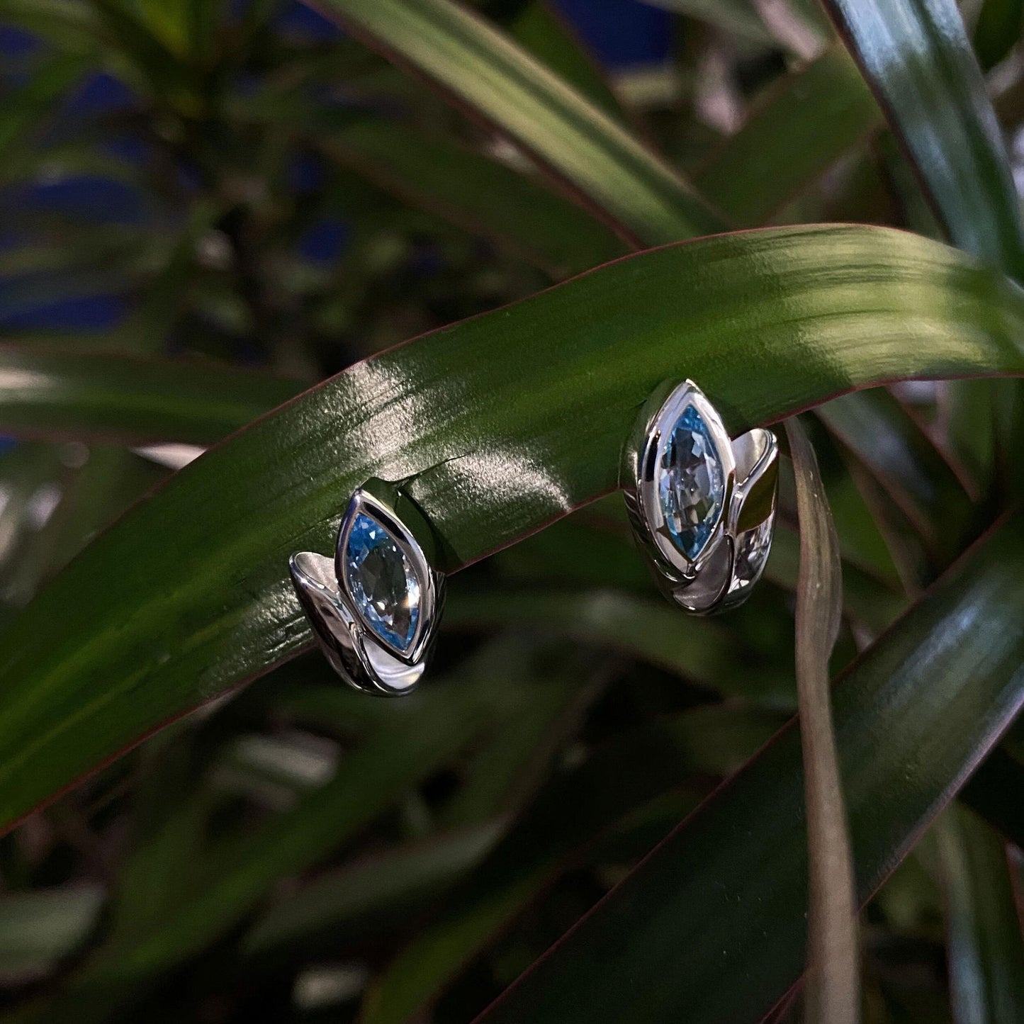 Photo of Blue Topaz Marquise Huggies in a decorative scene, designed by , available at Plumb Gold & Plumb Silver