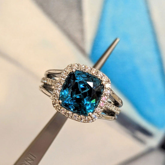 Photo of Blue Zircon Cushion Halo Ring in a decorative scene, designed by , available at Plumb Gold & Plumb Silver